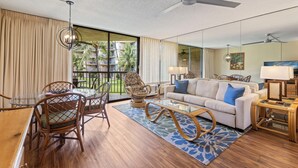 A modern living room with a glass dining table, wicker chairs, a sofa with blue cushions, and a ceiling fan. - Maui Sunset Vacation Rentals B206 (Kihei)