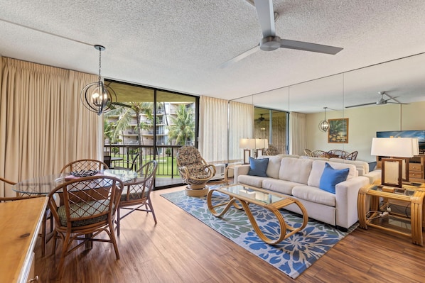 A modern living room with a glass dining table, wicker chairs, a sofa with blue cushions, and a ceiling fan.