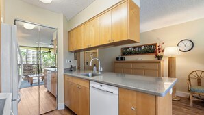 A modern kitchen with wooden cabinets, a dishwasher, a sink, and a dining area with a view of the outdoors. - Maui Sunset Vacation Rentals B206 (Kihei)