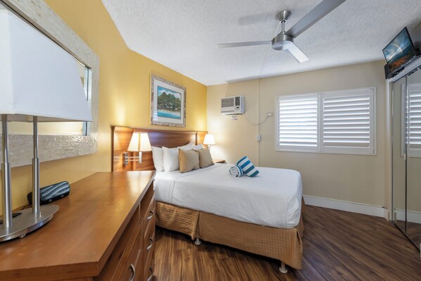 A hotel room with a bed, bedside table, lamp, ceiling fan, air conditioning unit, and a window with blinds. - Maui Sunset Vacation Rentals B206 (Kihei)