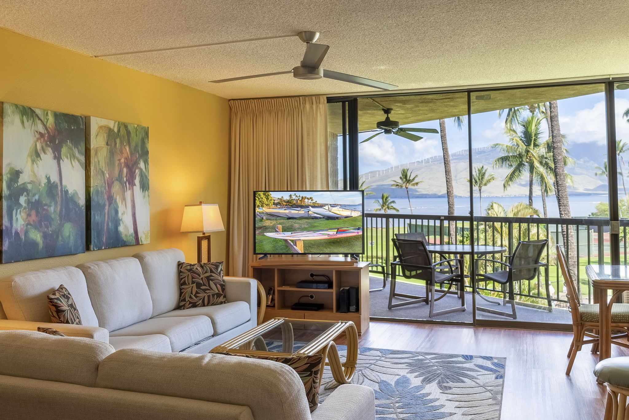 Een woonkamer met een bank, televisie en balkon met uitzicht op palmbomen en de oceaan.