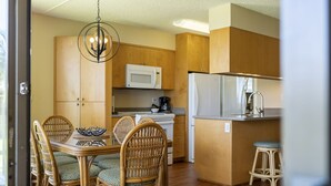 A compact kitchen with wooden cabinets, a microwave, a refrigerator, and a dining area with wicker chairs and a table. - Maui Sunset Vacation Rentals- B318 (Kihei)