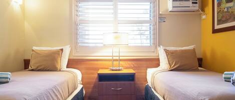 Et hotelværelse med to senge, et natbord med skuffer, en lampe, en aircondition og et vindue med persienner.