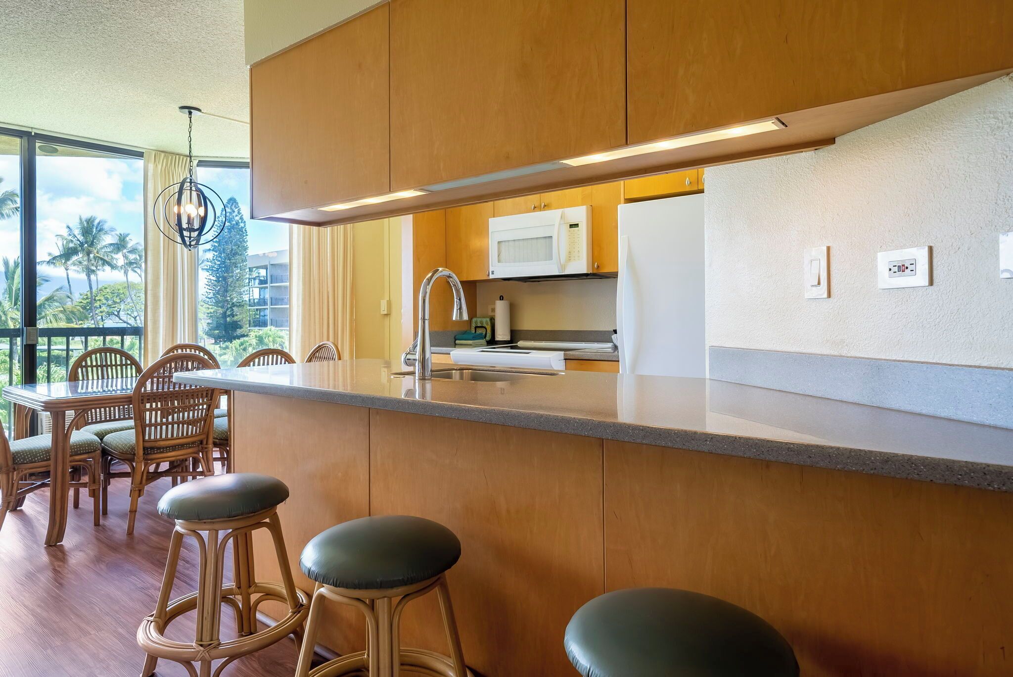 A modern kitchen with wooden cabinets, a stainless steel sink, and a white microwave. There are two bar stools and a dining table with chairs.
