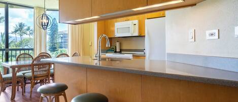 A modern kitchen with wooden cabinets, a stainless steel sink, and a white microwave. There are two bar stools and a dining table with chairs.