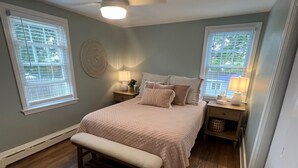 A bedroom with a bed, two bedside tables, a bench, and a wall-mounted fan. - Cheerful, Newly Renovated Cottage close to Newport w/ hot tub! (MIddletown)