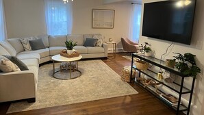 A modern living room with a sectional sofa, a round coffee table, a TV mounted on the wall, and a ceiling fan. - Cheerful, Newly Renovated Cottage close to Newport w/ hot tub! (MIddletown)
