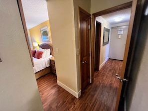 A narrow hallway with wooden flooring leading to a bedroom with a bed and nightstands.