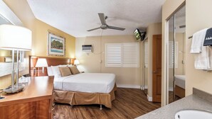 A hotel room with a bed, a desk with a lamp, a ceiling fan, a painting on the wall, and a bathroom with a sink and towel rack. - Maui Sunset Vacation Rentals B406 (Kihei)