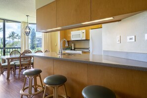 Eine moderne Küche mit Holzschränken, einer Edelstahlspüle und einer weißen Mikrowelle. Zwei Barhocker sowie ein Esstisch mit Stühlen sind ebenfalls vorhanden.