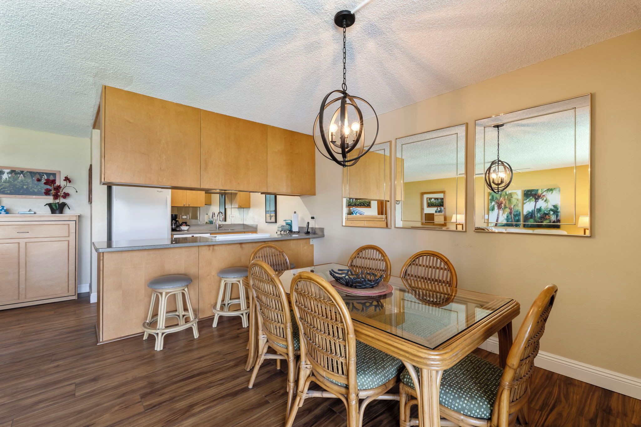 A modern kitchen with wooden cabinets, a glass dining table, wicker chairs, and a hanging light fixture.