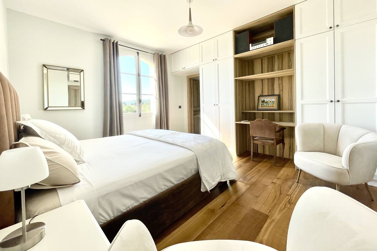 A modern bedroom with a large bed, a desk, and a chair. There is a window with curtains, a ceiling light, and built-in shelves.