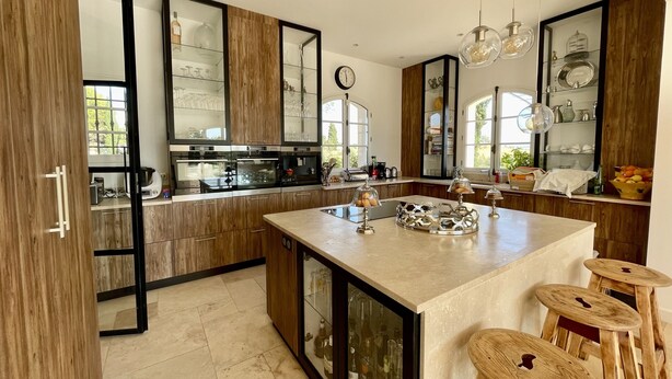 A modern kitchen with wooden cabinets, a central island, bar stools, and a large window. - La Soleillade Aixoise (Aix-en-Provence)