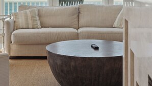 Un salon moderne avec un canapé beige, une table basse ronde et une vue sur l’océan grâce à de grandes fenêtres.