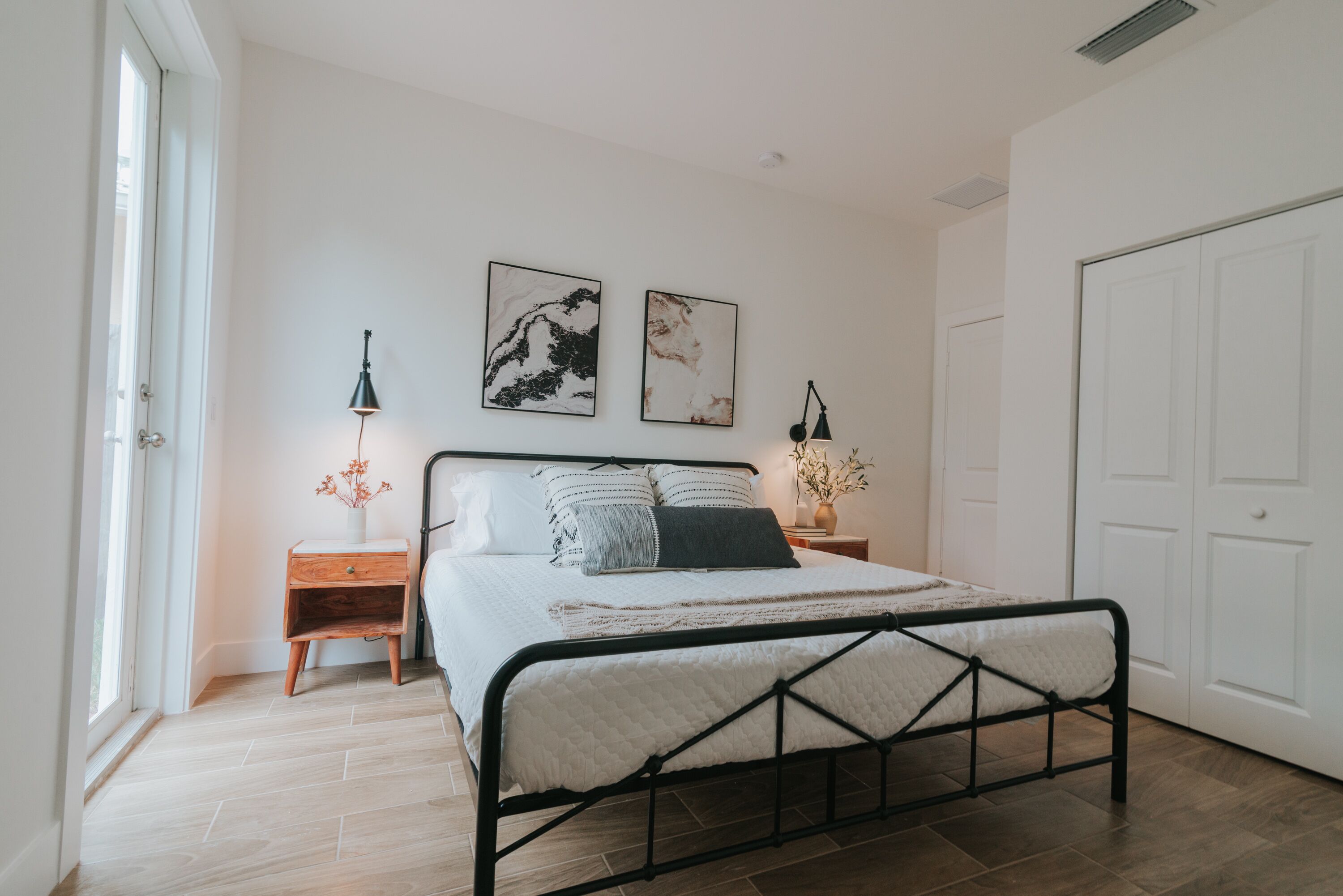 A bedroom with a bed, two bedside tables, a nightstand, a door, and two wall-mounted pictures.