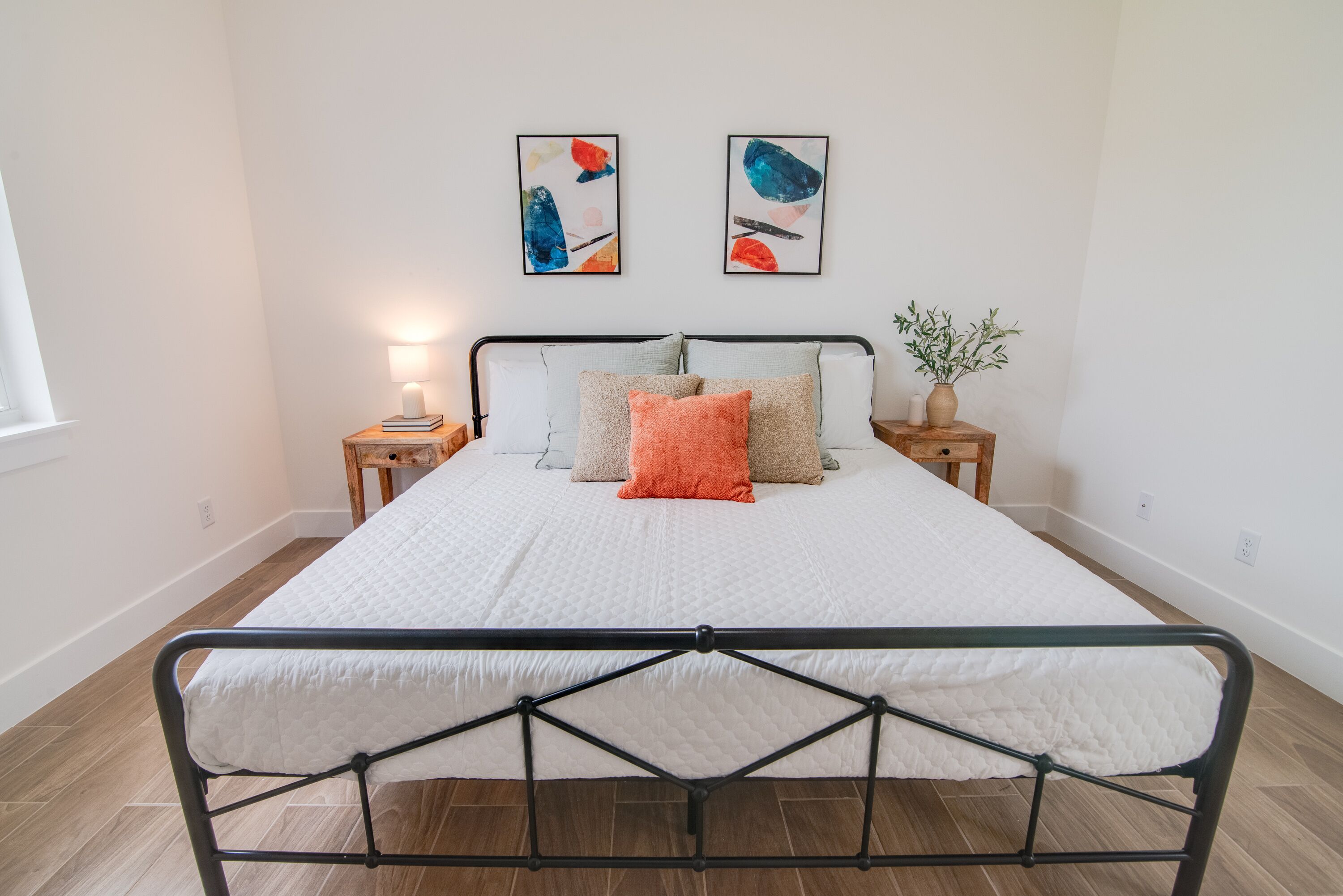A bedroom with a bed, two bedside tables, a nightstand, a floor lamp, and two abstract paintings on the wall.