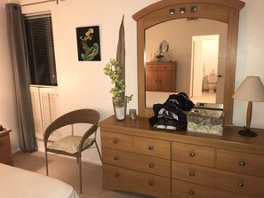 A bedroom with a wooden dresser, a chair, a bed, a mirror, and a window with blinds.