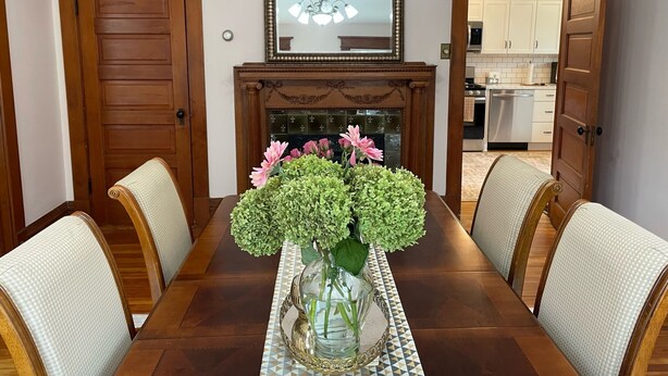 Une salle à manger avec une table en bois, des chaises, un vase de fleurs et une cheminée.