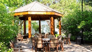 A wooden gazebo with a tiled roof surrounded by outdoor furniture, set in a lush garden.