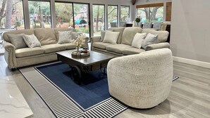 Ein geräumiges Wohnzimmer mit großen Fenstern, einem Ecksofa, einem Couchtisch und einem gemusterten Sessel.