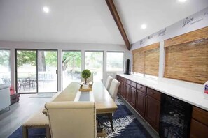 A modern kitchen with a long dining table, wooden cabinets, and a large window providing a view of the outdoors.