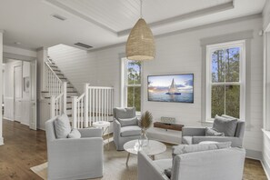A modern living room with a staircase, a large window with a view of the sea, and a flat-screen TV mounted on the wall.