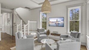 A modern living room with a staircase, a large window with a view of the sea, and a flat-screen TV mounted on the wall. - Beargrass. 4 Bedroom Newly Updated with 6-Seater Golf Cart & Bikes! (Santa Rosa Beach)