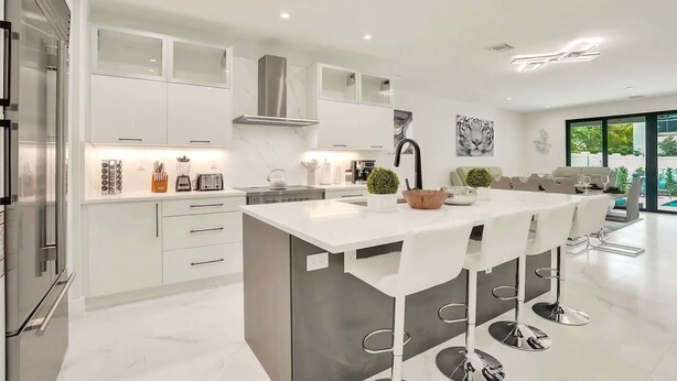 A modern kitchen with white cabinetry, a central island with bar stools, and a large window offering a view of the outdoors.