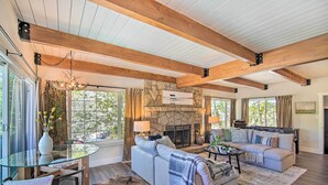 A spacious living room with a stone fireplace, wooden beams, and large windows. - Chic Lake House w/ Furnished Deck & Hot Tub! (Lake Arrowhead)