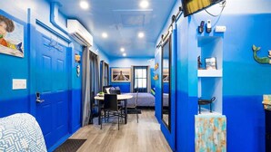 A room with blue walls, a wooden floor, a bed, a desk with a chair, and a TV mounted on the wall. - Experience Tiny House Weddings & Retreats (Cape Charles)