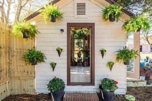 A small white building with a brown door, surrounded by a wooden fence. There are several potted plants both hanging and placed on the ground.