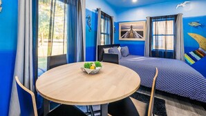 A room with a bed, a round wooden table with a bowl of fruit, and a window with curtains. - Experience Tiny House Weddings & Retreats (Cape Charles)