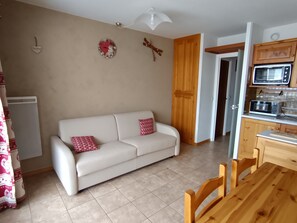 Ein Wohnzimmer mit einem weißen Sofa, roten Kissen mit Muster, einem Holzespeisetisch und einer Küchenzeile inklusive Mikrowelle und Schränken.