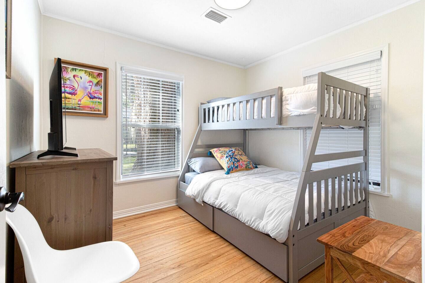 A bunk bed room with a desk and chair, a painting on the wall, and windows with blinds.