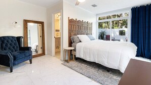 Une chambre à coucher comprenant un lit, une chaise, un ventilateur de plafond, une fenêtre avec des rideaux et une porte donnant sur une salle de bain.