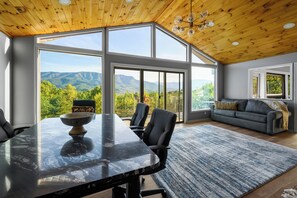Relax in a spacious living area with a large table and comfortable seating, featuring expansive doors with a view of the surrounding mountains.