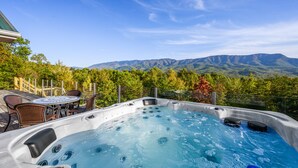 A hot tub with a mountain view, surrounded by a deck with chairs and a table.