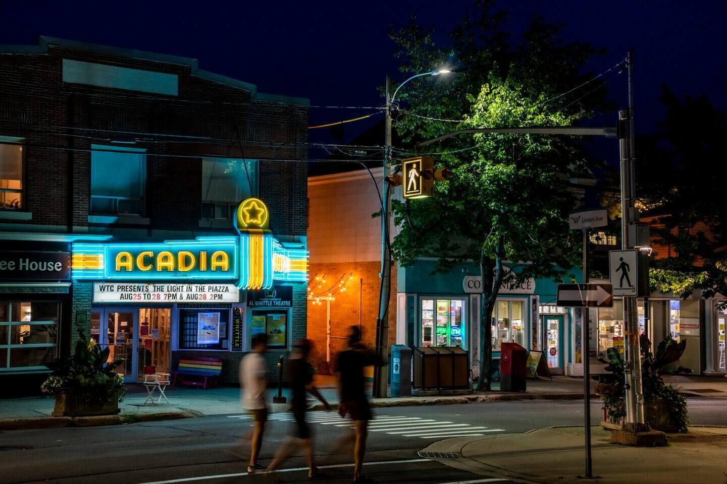 Escena nocturna en la calle con un brillante cartel luminoso del teatro ACADIA, un paso de peatones y transeúntes.