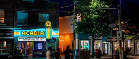 Escena nocturna en la calle con un brillante cartel luminoso del teatro ACADIA, un paso de peatones y transeúntes.