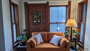 A cozy living room with a brown sofa, two pillows, a framed botanical print on the wall, a lamp on a side table, and a potted plant. - Flamingo Suite (Downtown Wolfville) (Wolfville)