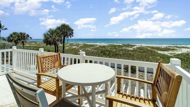 A balcony with a white railing, wooden chairs, and a small round table. - Sea Watch: 4B/4B Direct Oceanfront Home with Pool Near Downtown Cocoa Beach (Cocoa Beach)