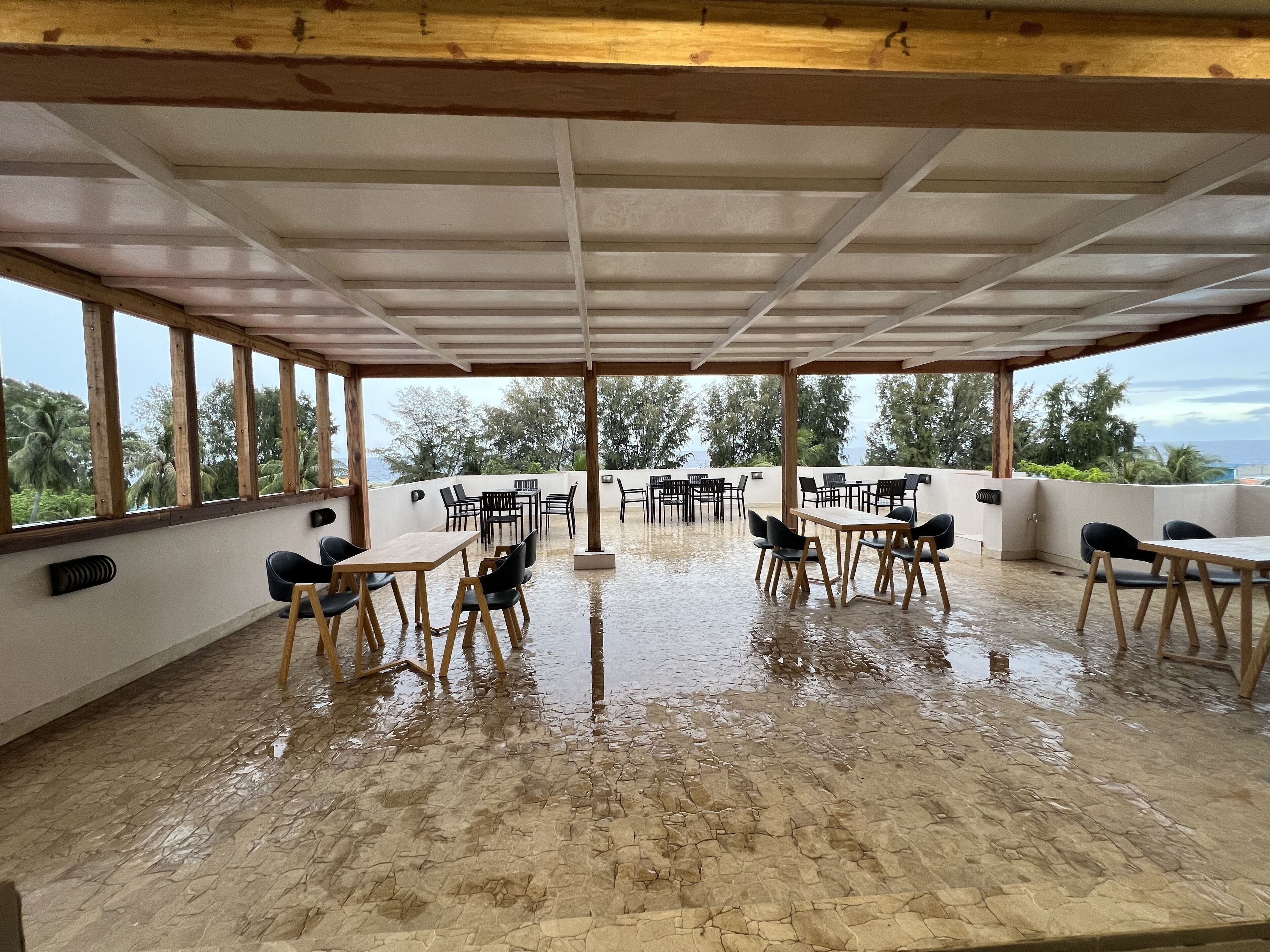 A covered outdoor seating area with tables and chairs, large windows, and a view of the sea.
