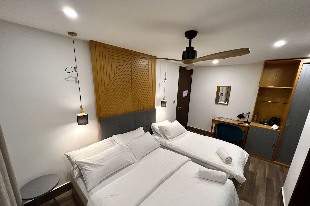 A hotel room with two beds, a wooden headboard, a ceiling fan, a desk with a chair, a mirror, and a small kitchenette.