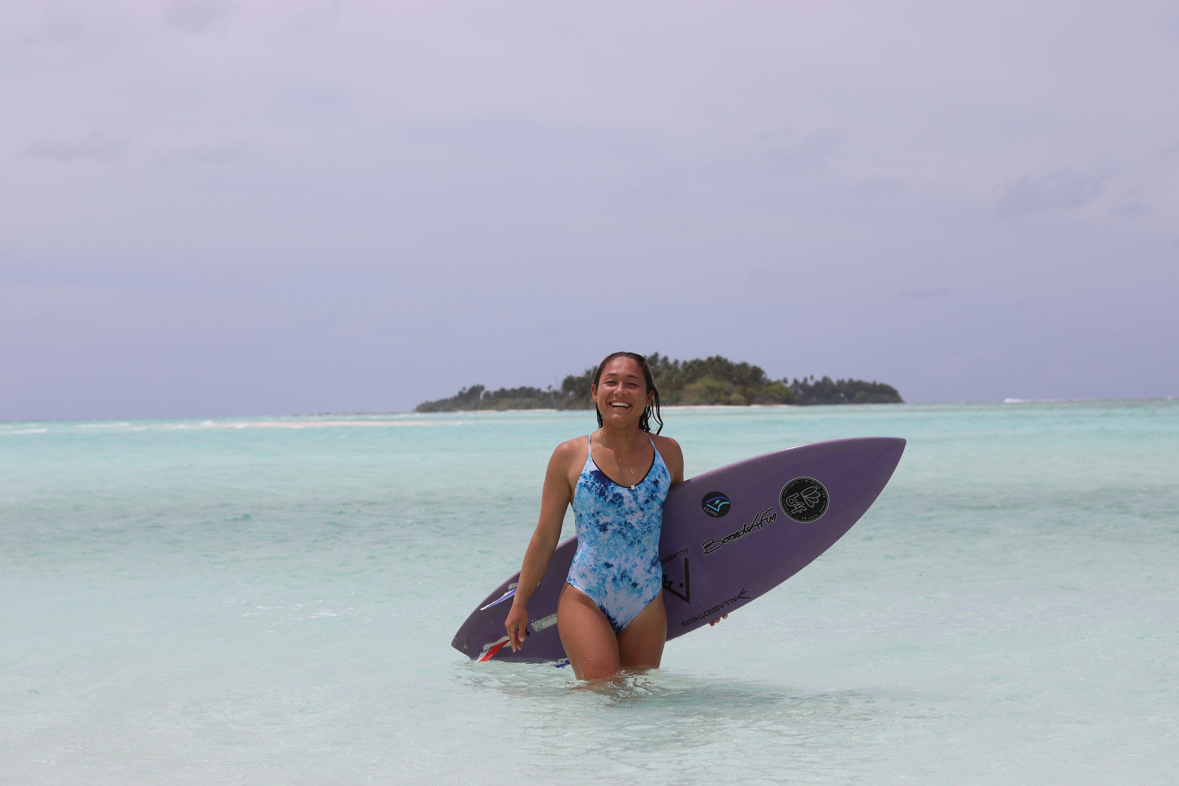 A person in a blue swimsuit holding a purple surfboard in clear water with a distant island.