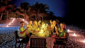 A group of people dining at an outdoor table with candles, palm trees, and a beach in the background. - Stay Mikado (Thimarafushi)