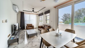 A modern living room with a ceiling fan, a flat-screen TV, leather sofas, a dining table, and large windows offering a view of the outdoors. - Stay Mikado (Thimarafushi)