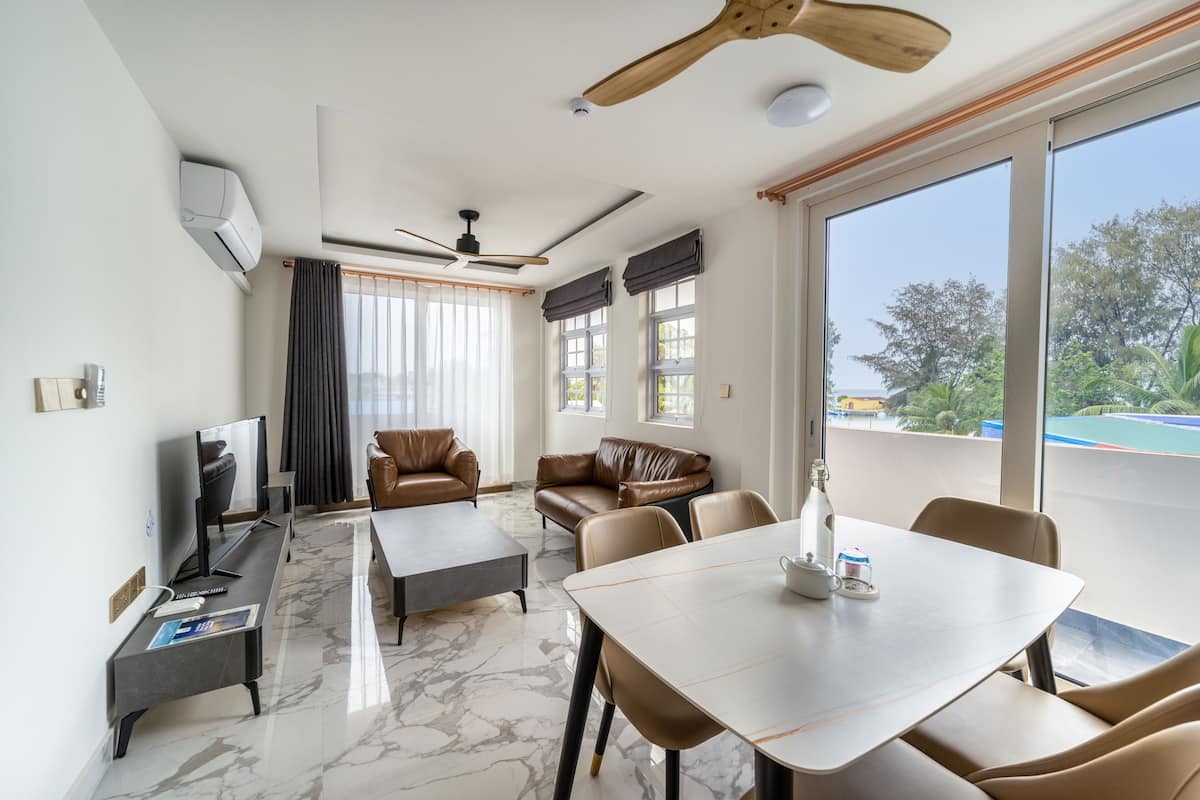 A modern living room with a ceiling fan, a flat-screen TV, leather sofas, a dining table, and large windows offering a view of the outdoors.