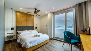 A modern hotel room with a large bed, a desk, a chair, a ceiling fan, and a view of the city through the window. - Stay Mikado (Thimarafushi)