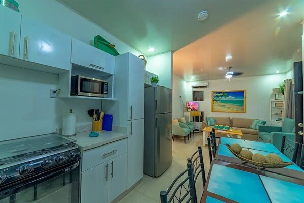 A modern kitchen with a gas stove, microwave, and refrigerator. There is a dining area with a table and chairs, and a living room with a sofa and a painting on the wall.