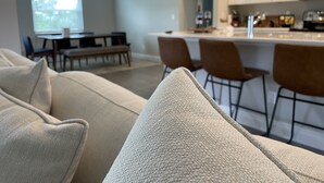 Una sala de estar moderna con un sofá color beige, un comedor con mesa y sillas, y una cocina con armarios blancos y una barra.